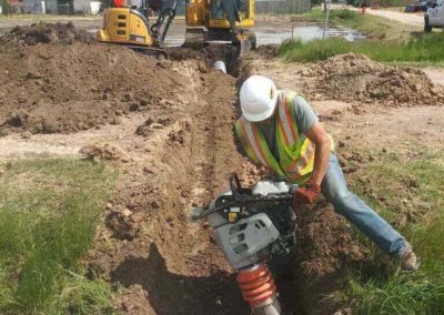 Construction worker working on a trench with equipment with a tractor in the background.